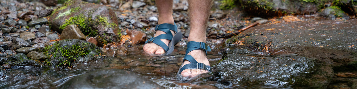 Man wearing blue Redway Sandals stepping into a rocky stream with mossy rocks nearby