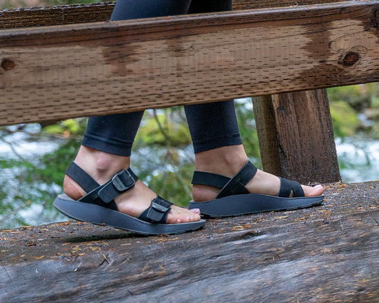 Woman wearing black leggings and black Salinas Leather Sandals walking across a wooden bridge in the forest